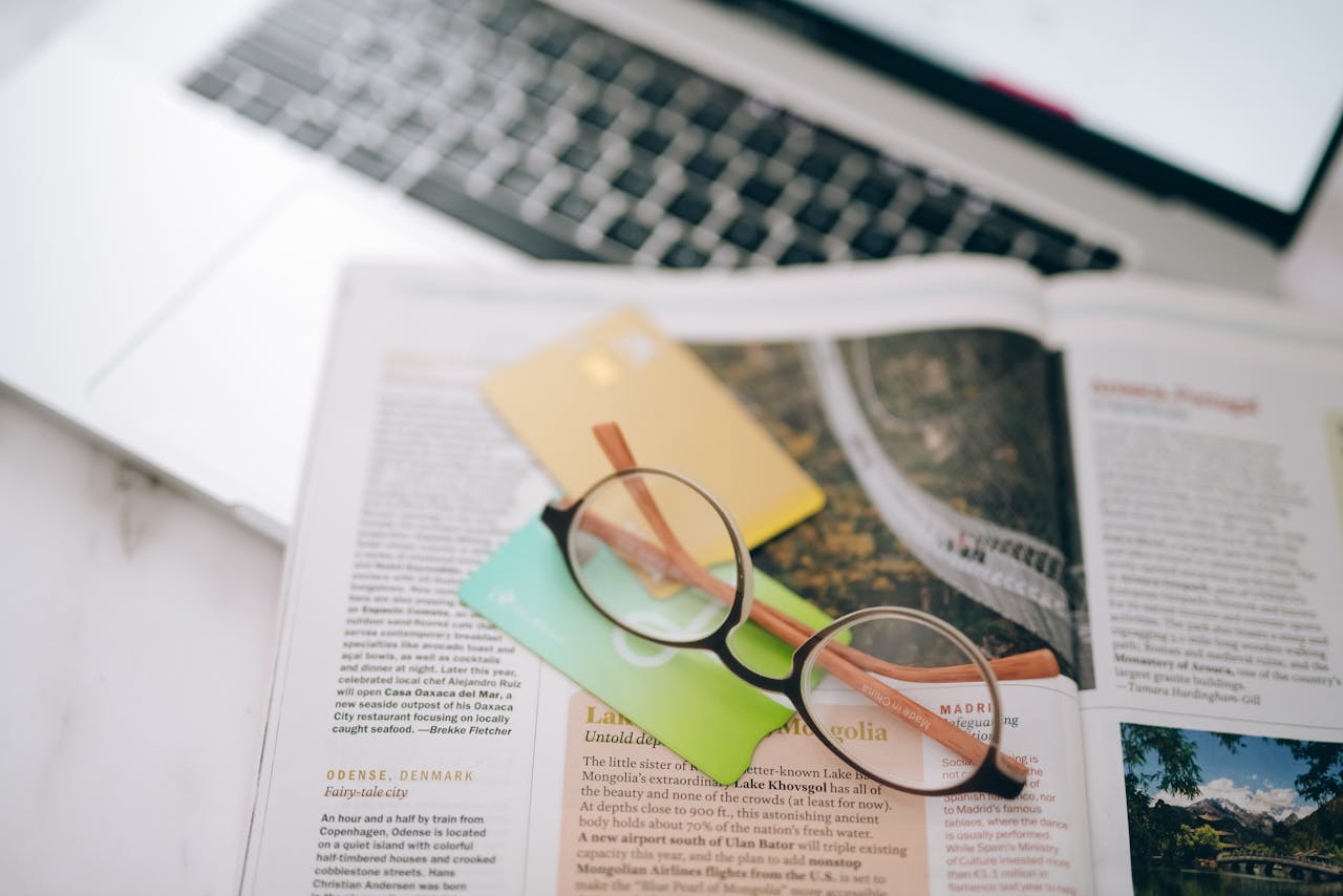 about-04 Overhead view of reading glasses, credit cards, and a magazine with a blurred laptop in the background.
